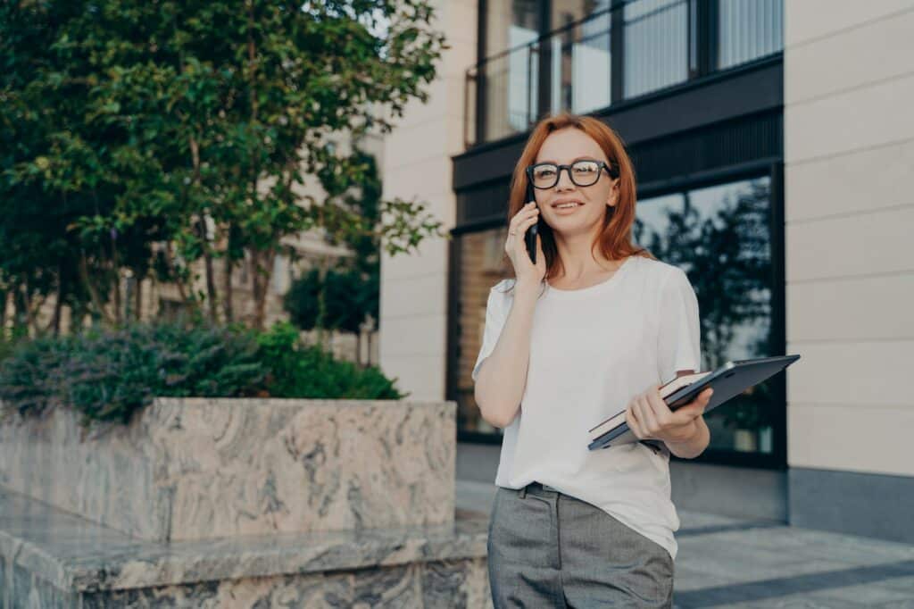 Young smiling european businesswoman calling on smartphone while walking in city with laptop in hand